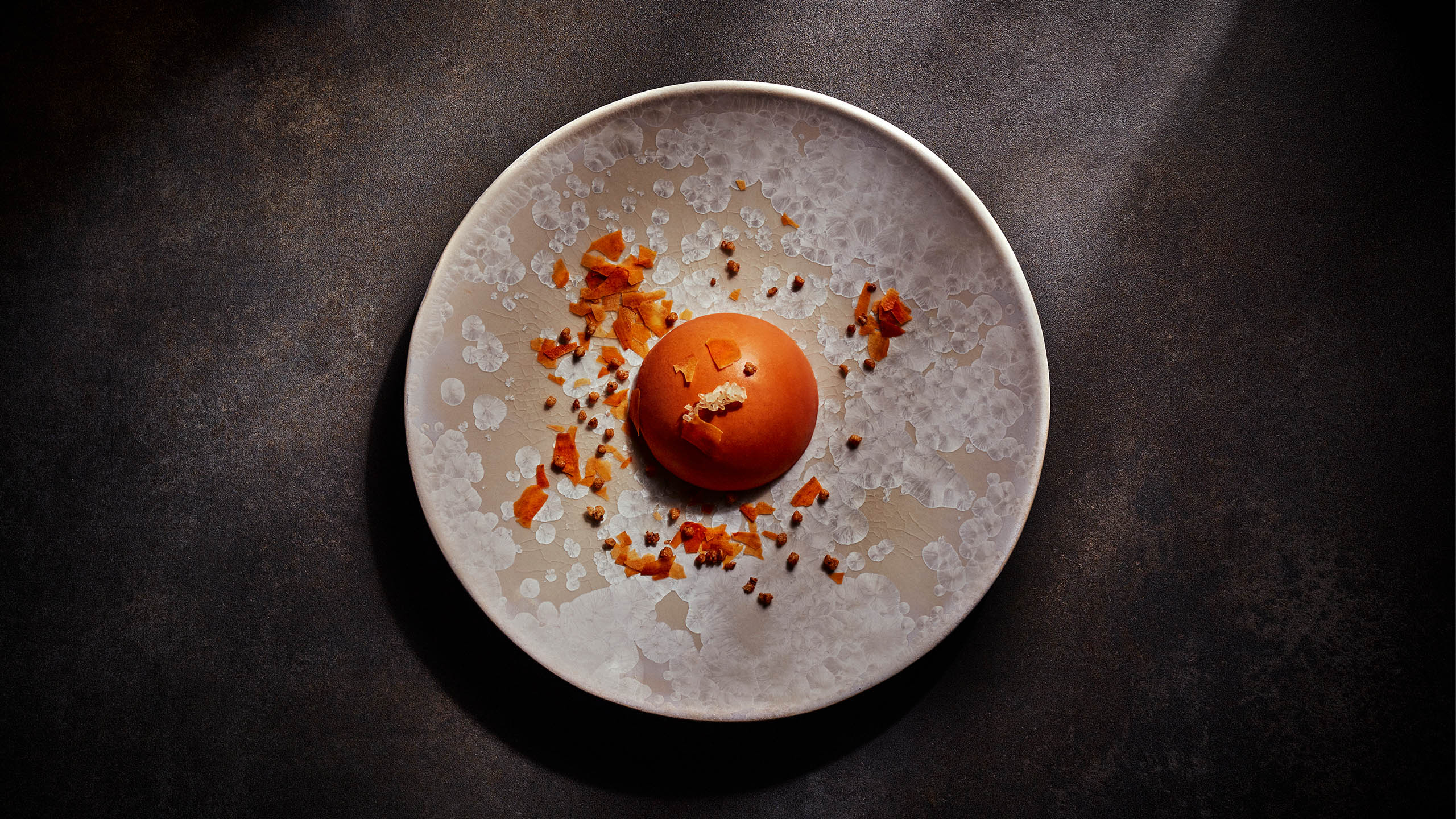 eine orangene Halbkugel liegt als Dessert auf einem hell-gefleckten Keramikteller