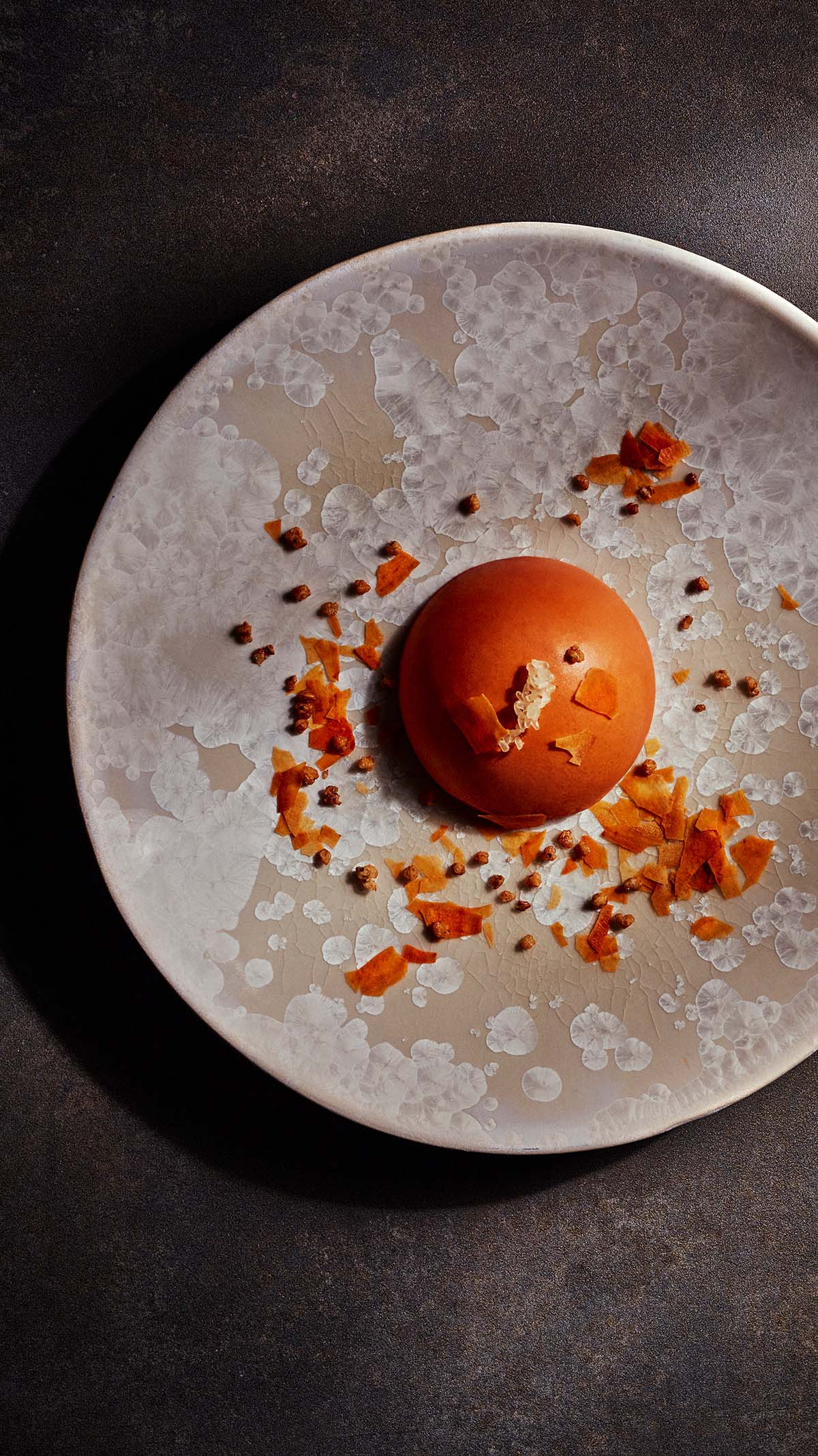 eine orangene Halbkugel liegt als Dessert auf einem hell-gefleckten Keramikteller
