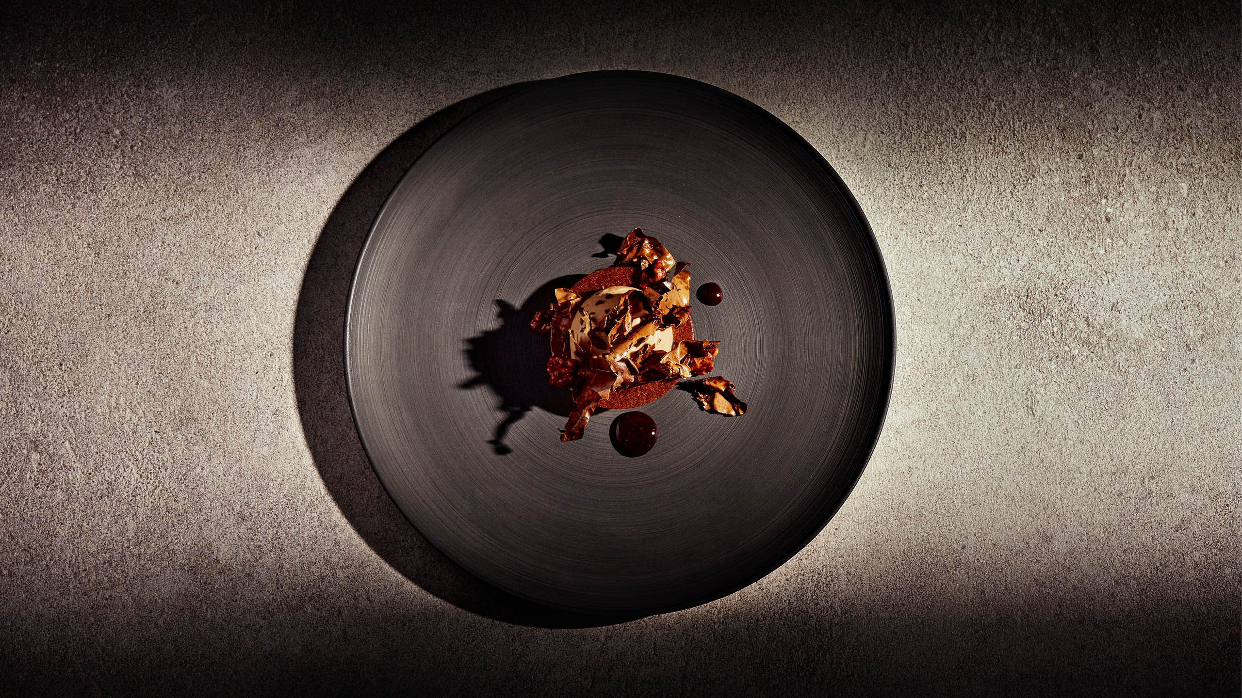 auf dunklem Teller zeigt sich ein bräunliches Dessert mit Creme und zwei Klecksen